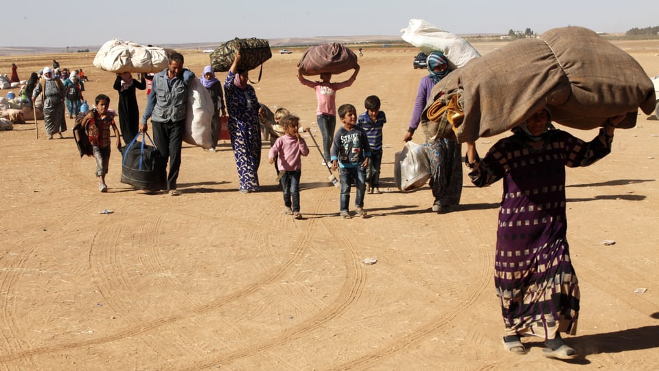 Syrische Familien tragen grosse Pakete auf dem Kopf und ziehen über eine Landstrasse.