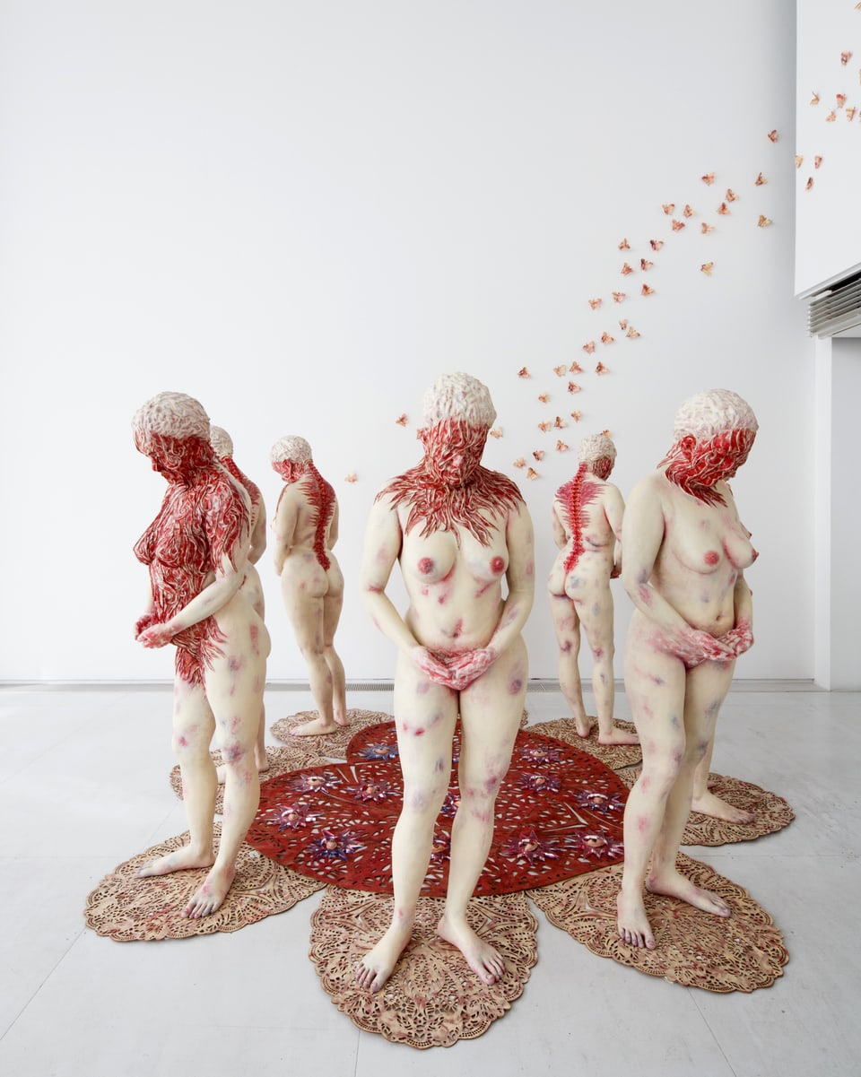 8 Frauenskulpturen in einem Kreis stehend, mit teilweise rot überwucherter Haut.