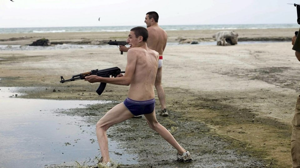 Zwei Jugendliche in Unterhosen schiessen am Strand mit Sturmgewehren.