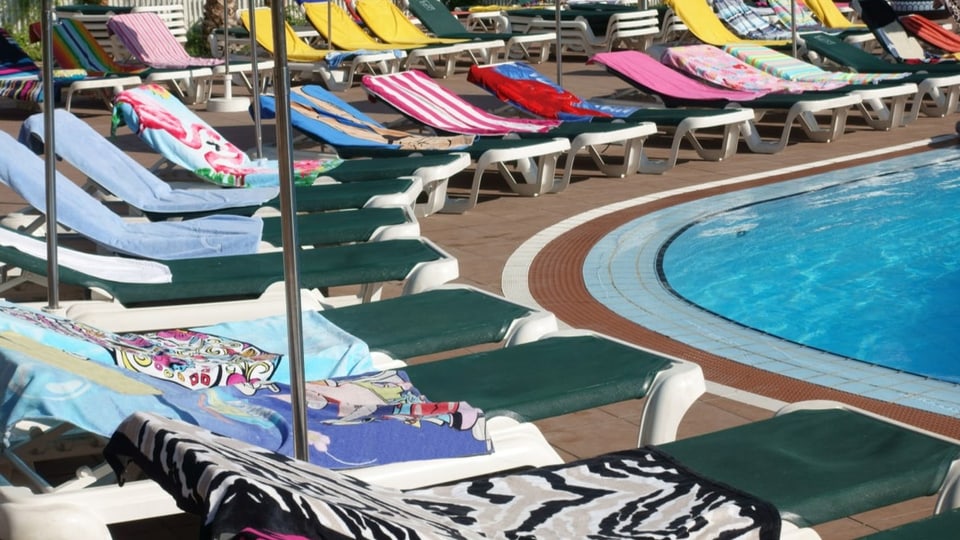 Foto von dunkelgrünen Liegestühlen im Halbkreis um Pool, alle mit bunten Tüchern besetzt, keine Menschen.