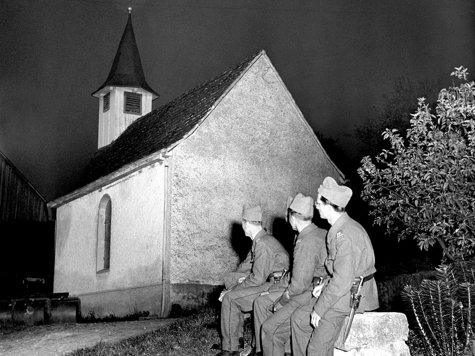 Drei Soldaten sitzen vor einer kleinen Dorfkirche.
