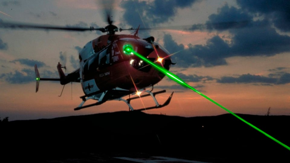 Ein Rega-Helikopter bei Dämmerung, auf den ein grüner Laserstrahl gerichtet ist (Photomontage).