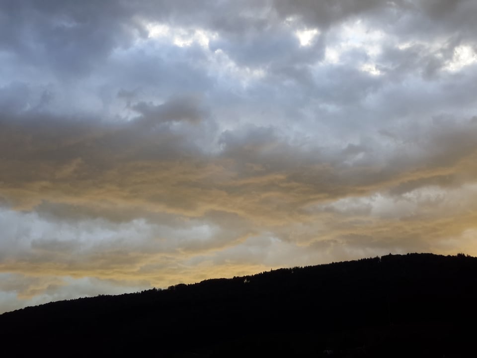 Grau-gelbes Wolkengewirr vor noch dunklem Hügel.