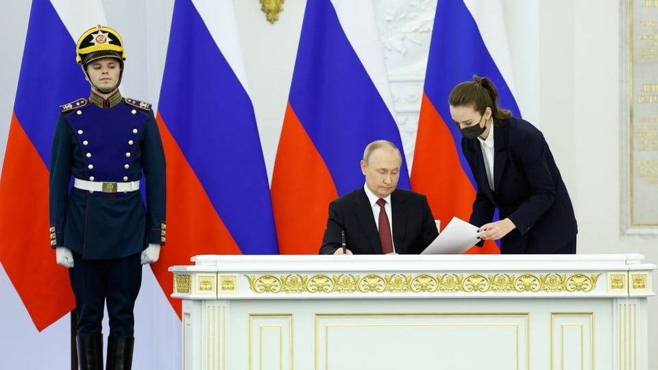 Putin am Pult Sitzend mit einem Stift in der Hand. Eine Frau reicht ihm ein Dokument zur Unterschrift.