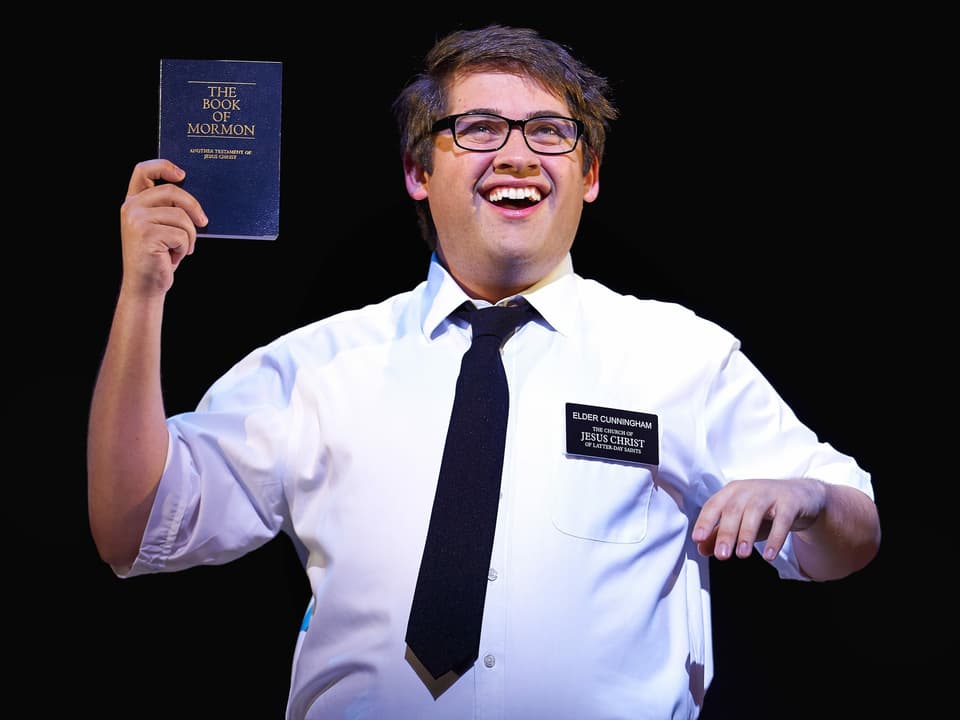 Ein Bild aus dem Musical "Book of Mormon". Ein Darsteller hält ein solches Buch hoch.