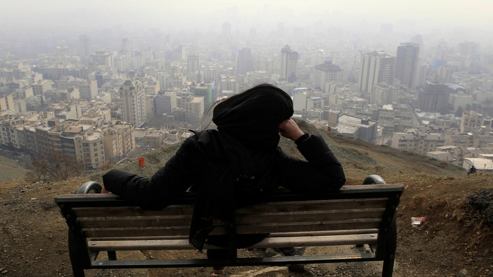 Frau mit Tschador sitzt auf Bank, im Hintergrund Teheran. 