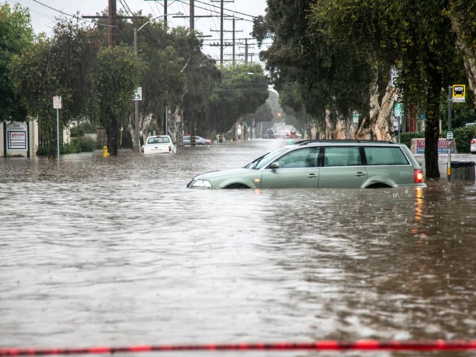 Eine von mediterranen Bäumen gesäumte Strasse steht unter Wasser, im Vordergrund steht ein verlassenes Auto.