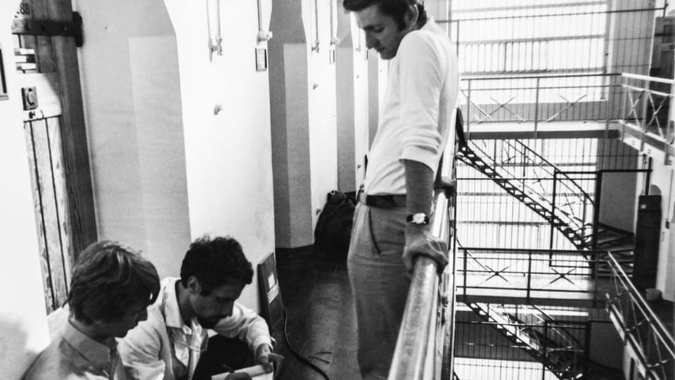 Schwarzweiss-Foto zwei junge Männer sitzen, einer lehnt stehend am Geländer. Im Hintergrund gitterartiges Treppenhaus.