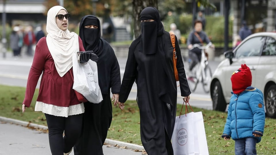 Zwei Frauen in der Mitte mit Burka, eine Frau links mit Hijab und ein kleiner Junge mit einer Kapuze spazieren.