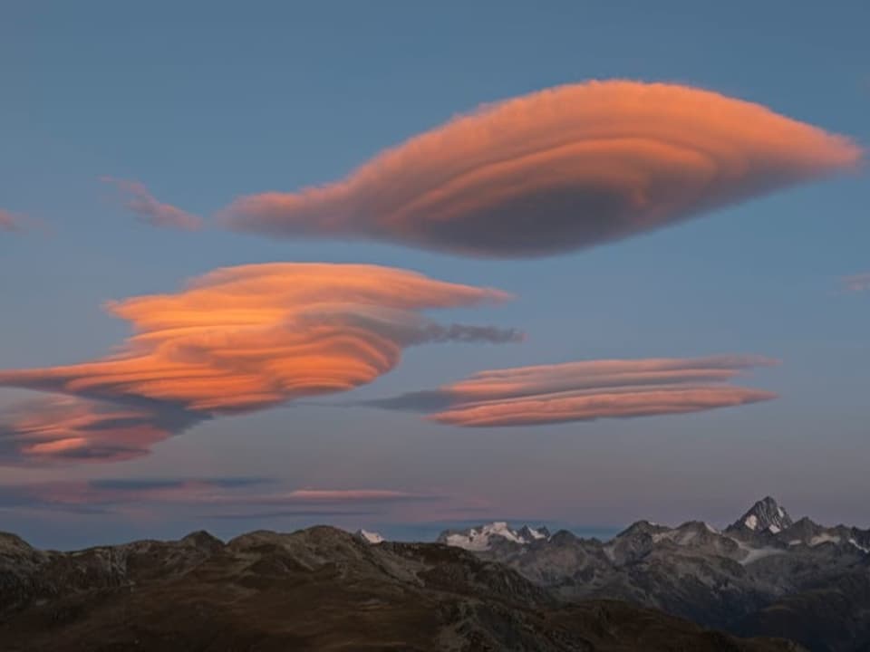 Alpenpanorame mit vielen Gipfeln, darüber linsenförmige Wolken, die rosa leuchten. 