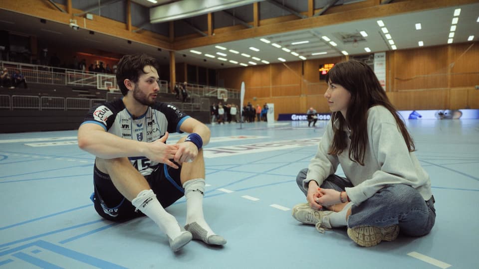 Ein Handballspieler und eine Frau sitzen auf dem Boden einer Turnhalle.