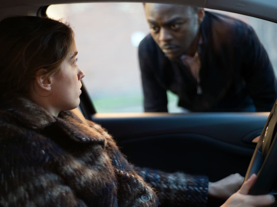 Eine Frau sitzt am Steuer eines Wagens, durch dessen Fenster ein Schwarzer sie anschaut.