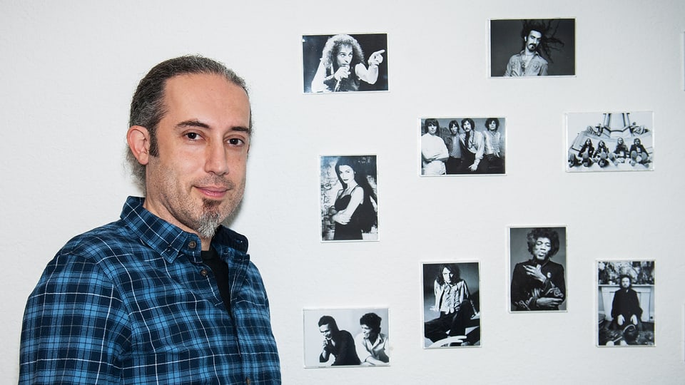 Ein Mann in kariertem Hemd steht vor einer Wand, an der mehrere Schwarz-Weiss-Bilder kleben.