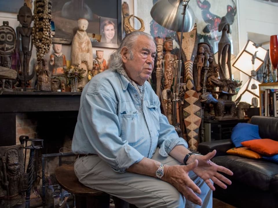 Ted Scapa in einem Zimmer mit zahlreichen Bildern und Statuen sitzend.