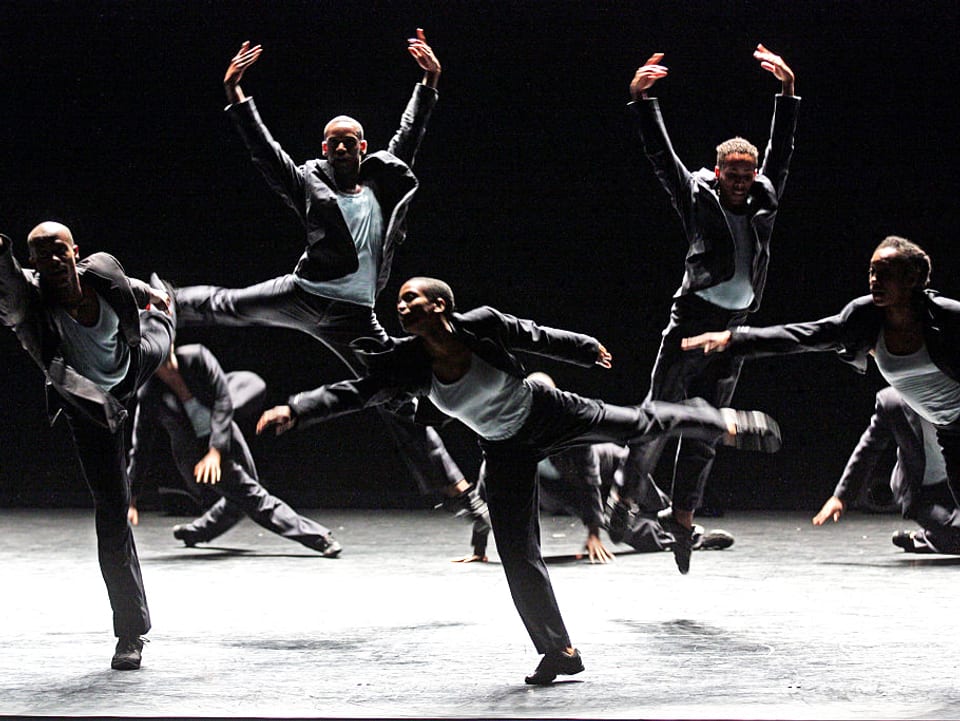 Männer springen in grauen Anzügen über die Bühne – in einer Mischung aus klassischem Ballett und zeitgenössischem Tanz.
