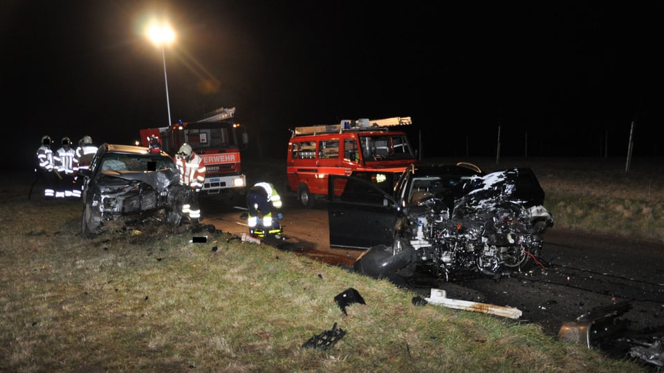 Zwei total zerstörte Fahrzeug, dahinter Einsatzfahrzeuge der Feuerwehr.