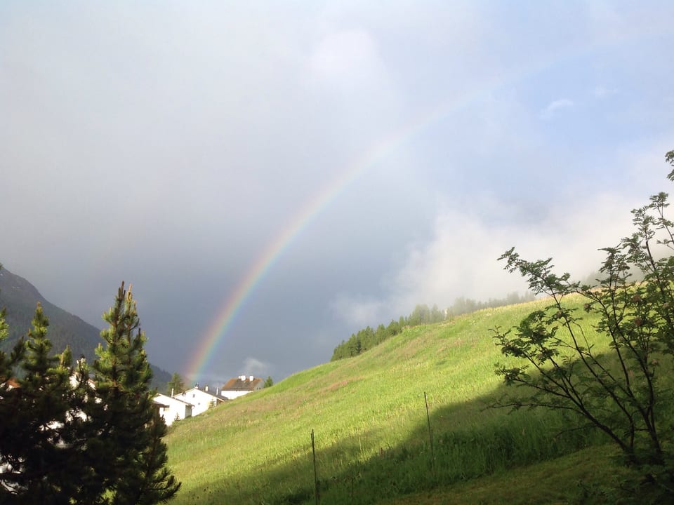 Über dem Tal im Hintergrund ragt ein Regenbogen in die Höhe, dahinter dunkle Regenwolken. Die Wiese im Vordergrund wird von der Sonne angestrahlt.