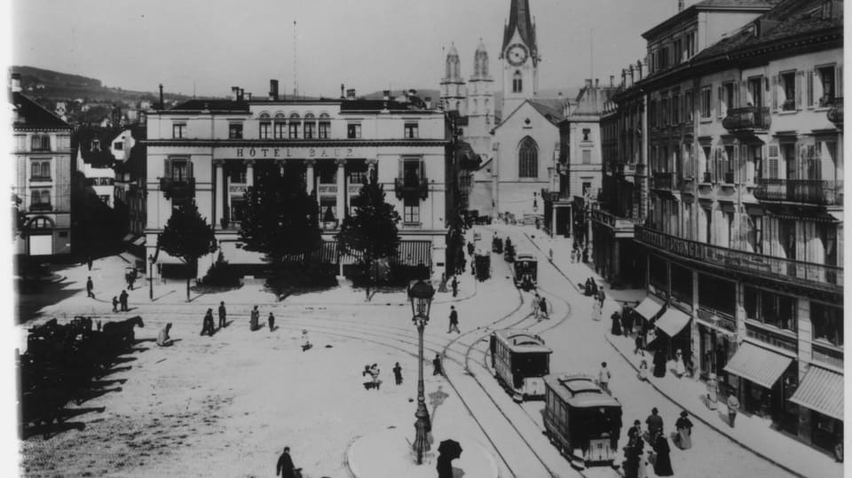 Historischer Platz mit Gebäuden, Menschen und Fahrzeugen.