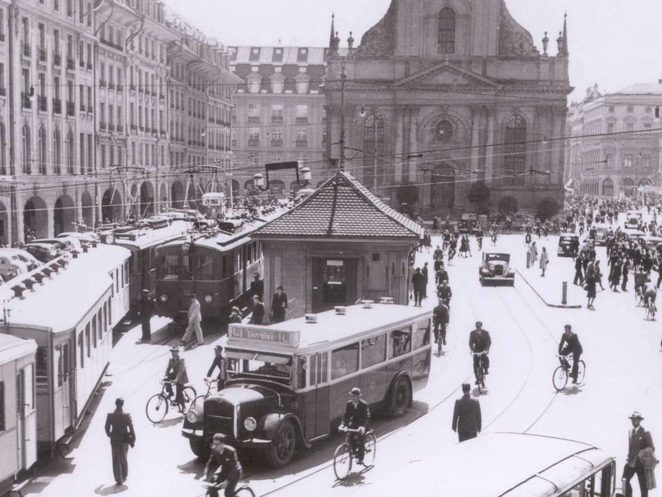 Schwarz-weiss-Bild: Im Hintergrund eine Kirche, vordran ein Postauto, links davon ein Zug, rechts davon Velofahrer und Fussgänger