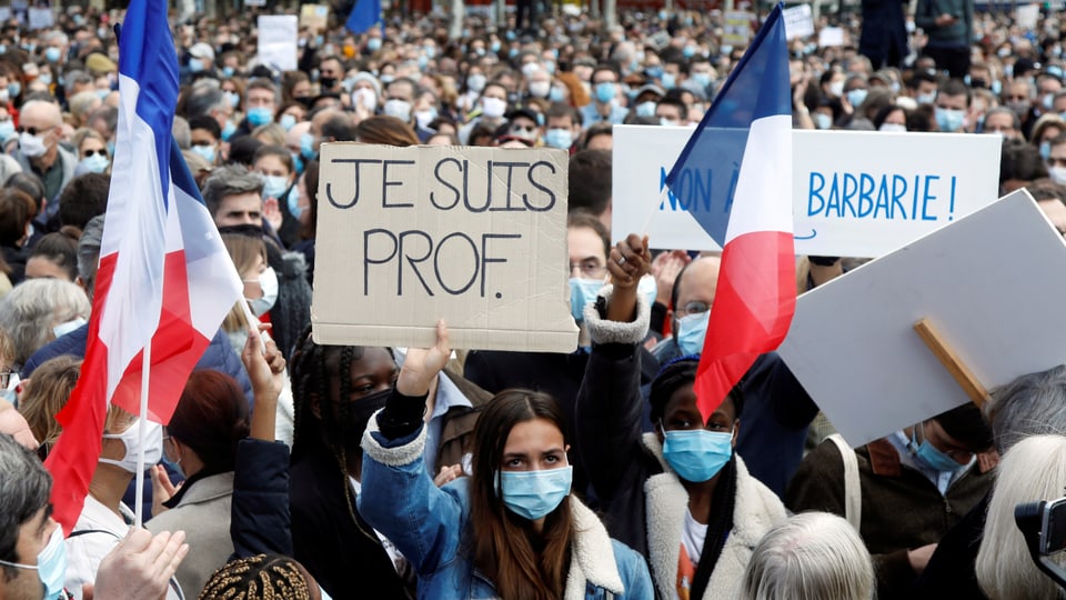 Eine Frau, die eine Gesichtsmaske trägt, hält ein Schild in die Luft, auf dem «Je suis prof.» steht.
