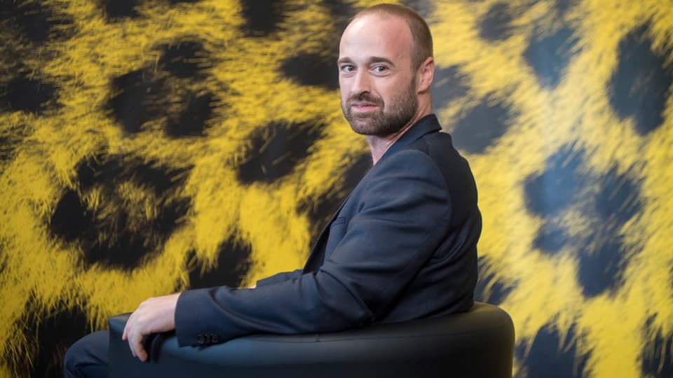 Das Bild zeigt Regisseur Dominik Locher vor einer gelb-schwarzen Wand. 
