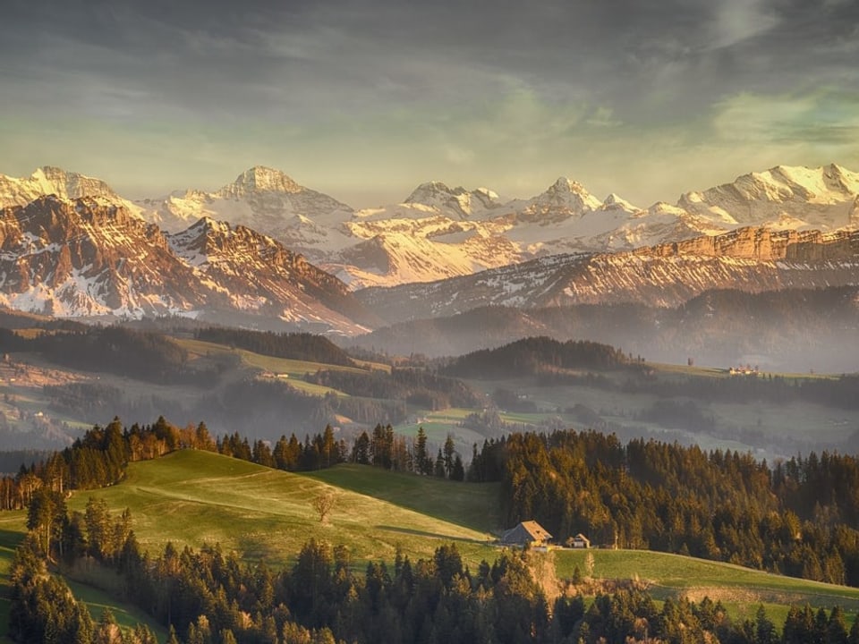 Blick über Felder, Wälder und Hügel auf die Schneeberge in der Ferne bei Sonnenuntergang