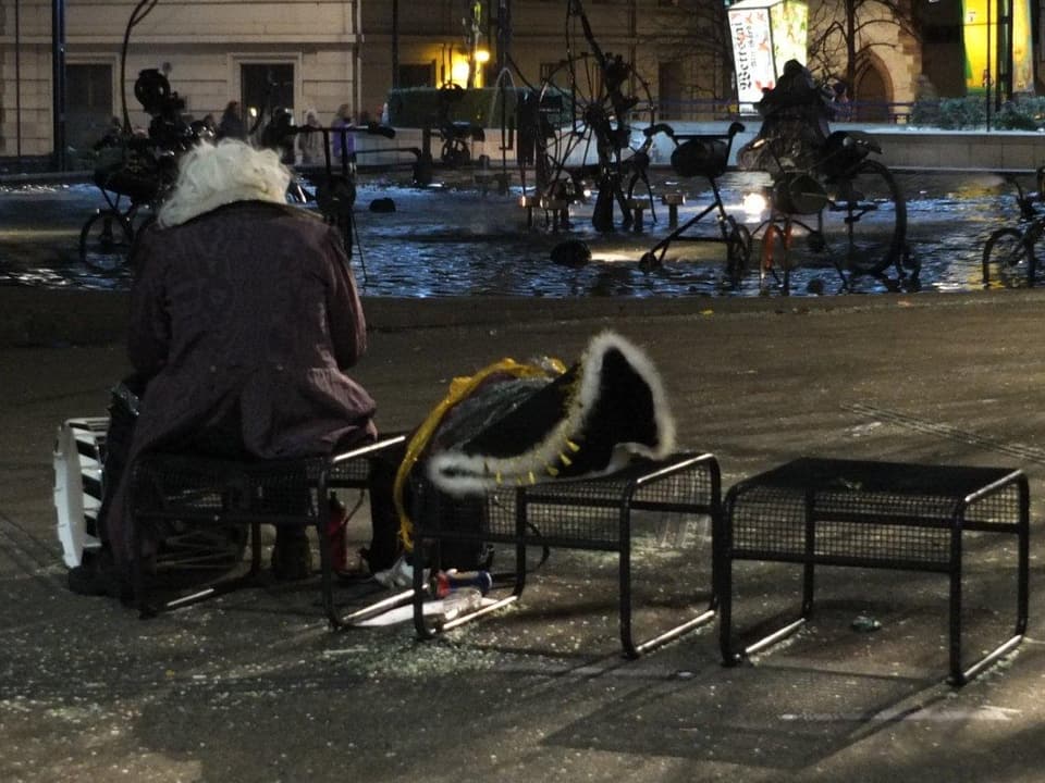 Kostümierter Fasnaächtler sitzt im Dunkeln auf einer Bank vor dem Tinguely-Brunnen.