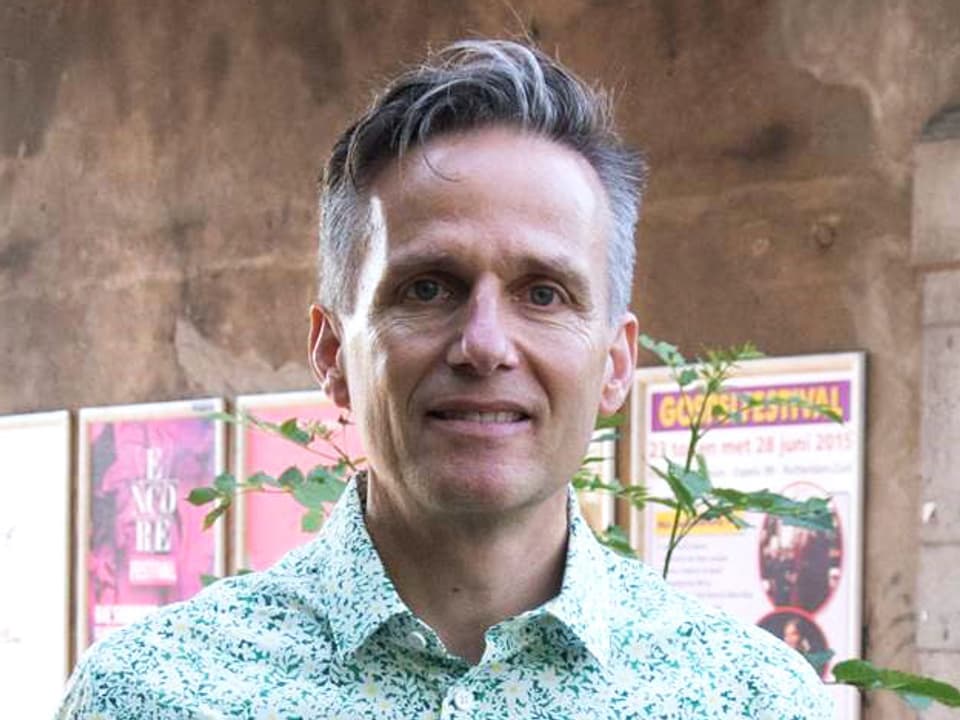 Christian Müller in grün-weiss-geblümbtem Hemd vor einer Mauer mit Plakaten