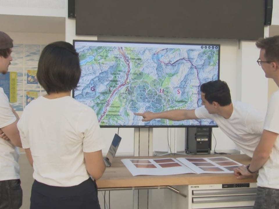 Vier Personen vor Bildschirm auf dem eine Karte zu sehen ist. Mann zeigt mit Finger auf Karte