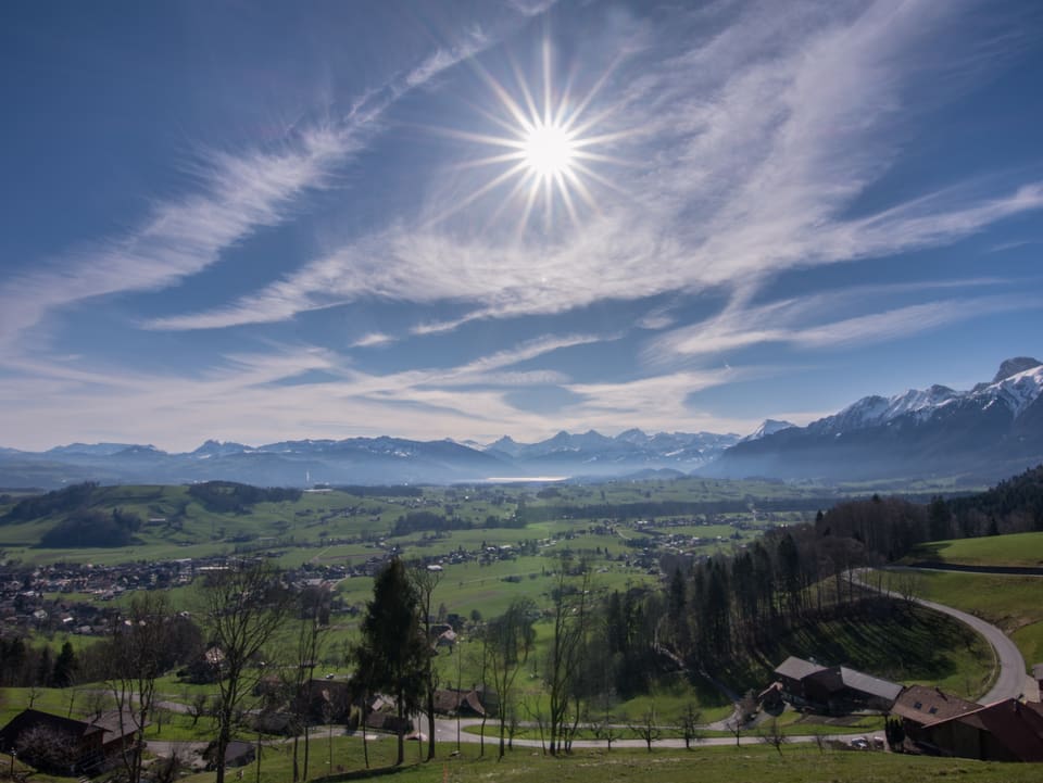 Die Sonne strahlt am Himmel, ein paar Wolkenschleier sind vorhanden, im Vordergrund  die grüne Landschaft und Häuser, dahinter die Alpen. 