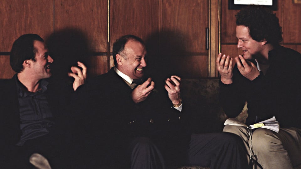 Drei Männer sitzen vor vor einer dunklen Holzwand und diskutieren.