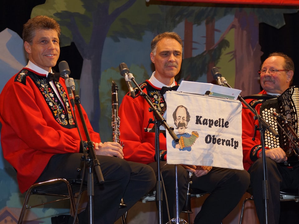 Drei Musikanten in roten Sennenkutten während einer Spielpause auf der Bühne.