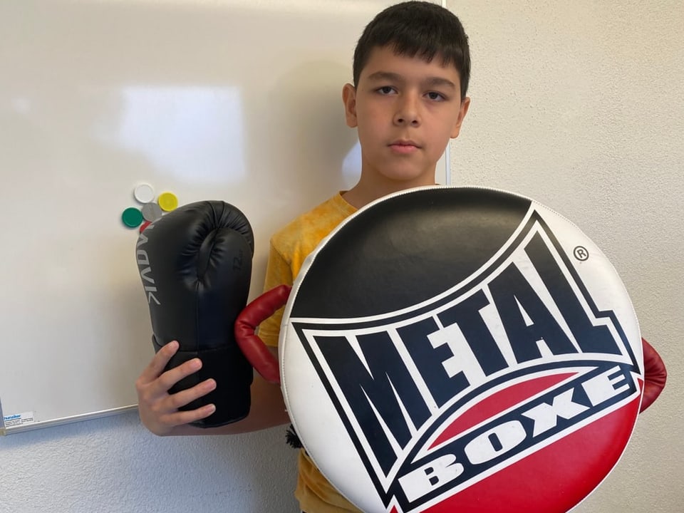 Ein Junge steht vor einer weissen Wand und hält einen Boxhandschuh und eine Kick Pratze