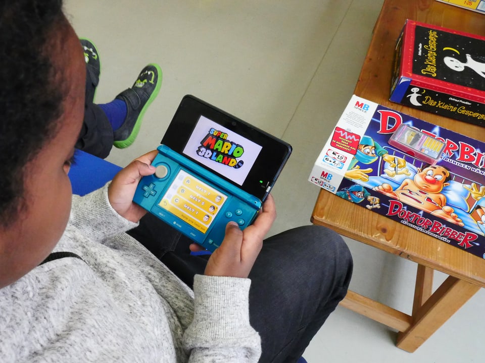Ein Kind spielt mit einer Nintendo-DS-Spielkonsole, im Hintergrund ist sind die Brettspiele «Dr. Bibber» und «Das kleine Gespenst» zu sehen.