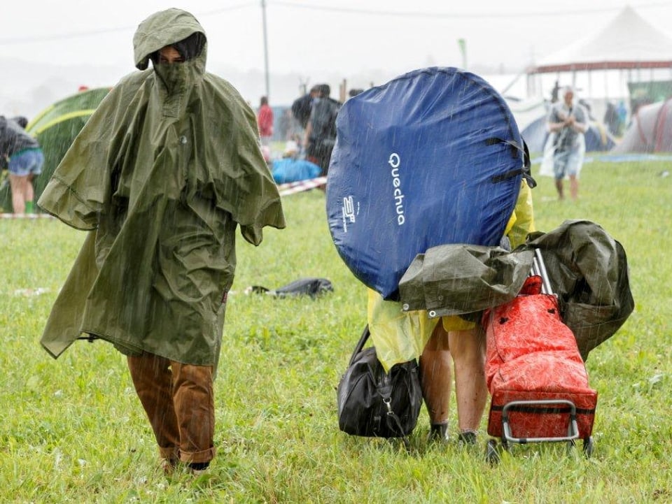 Links steht eine dick eingemummte Person. Nur noch die Stiefel und der Nasenspitz schauen aus dem Regenmantel hervor. Rechts im Bild eine Person von der man auch nur noch die Beine sieht. Die Person rechts ist allerdings von oben bis unten mit Campingartikel vollgeladen.