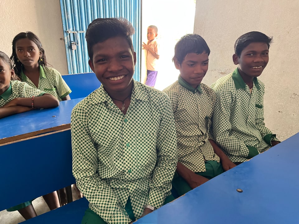 Santosh sitzt neben seinen Kollegen an einem Schulpult. Er lächelt in die Kamera.