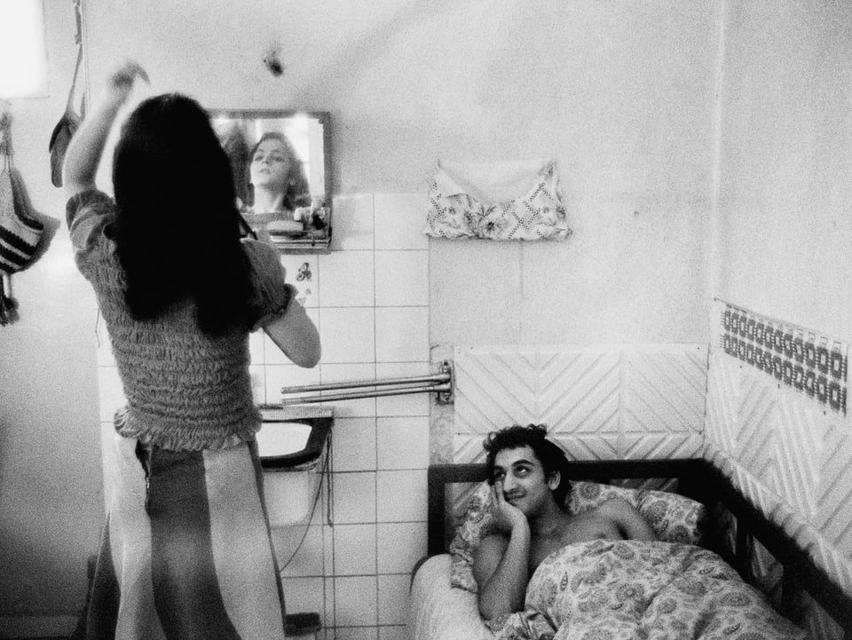 In einem kleinen Zimmer steht eine Frau vor dem Spiegel, ein Mann liegt daneben im Bett und schaut lächelnd zu.