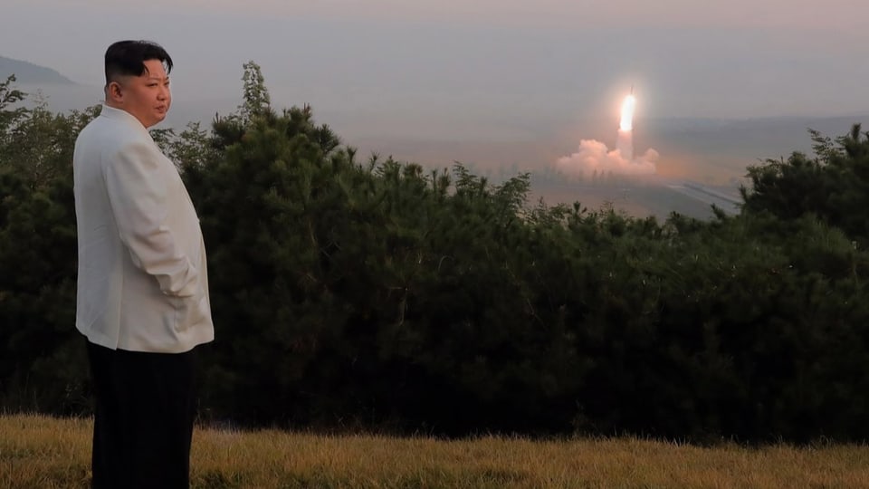 Kim Jong-un, schwarze Hose/helles Jackett, steht im Gras und schaut nach r., an einem Raketenlaunch in der Ferne vorbei.