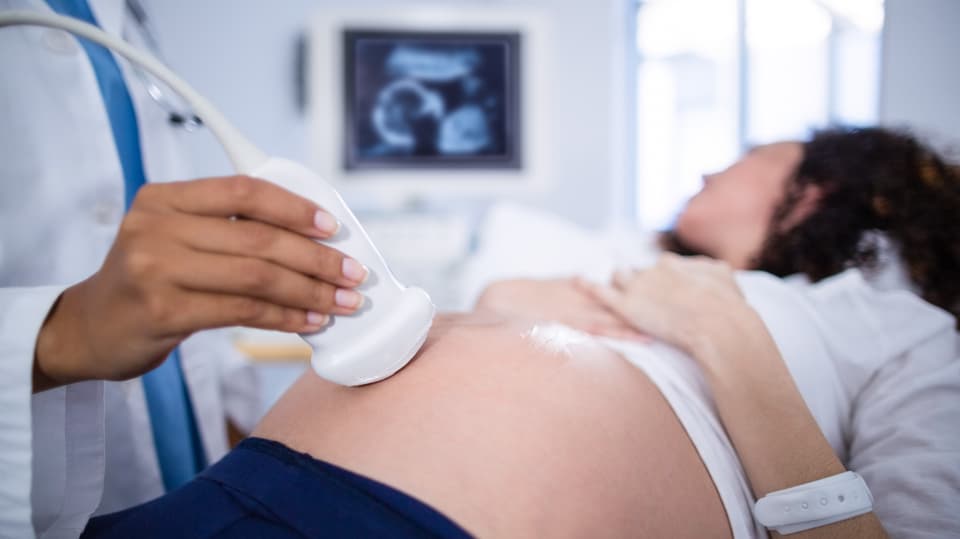 Eine Hand am Ultraschallgerät fährt den Bauch einer schwangeren Frau ab, die auf einen Monitor schaut. 