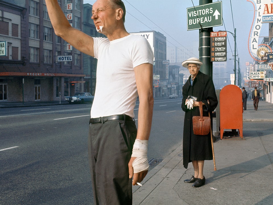 Ein Mann mit Verband an Kinn und Handgelenk hebt die Hand an der Strasse. Im Hintergrund eine alte Dame, die misstrauisch schaut. 