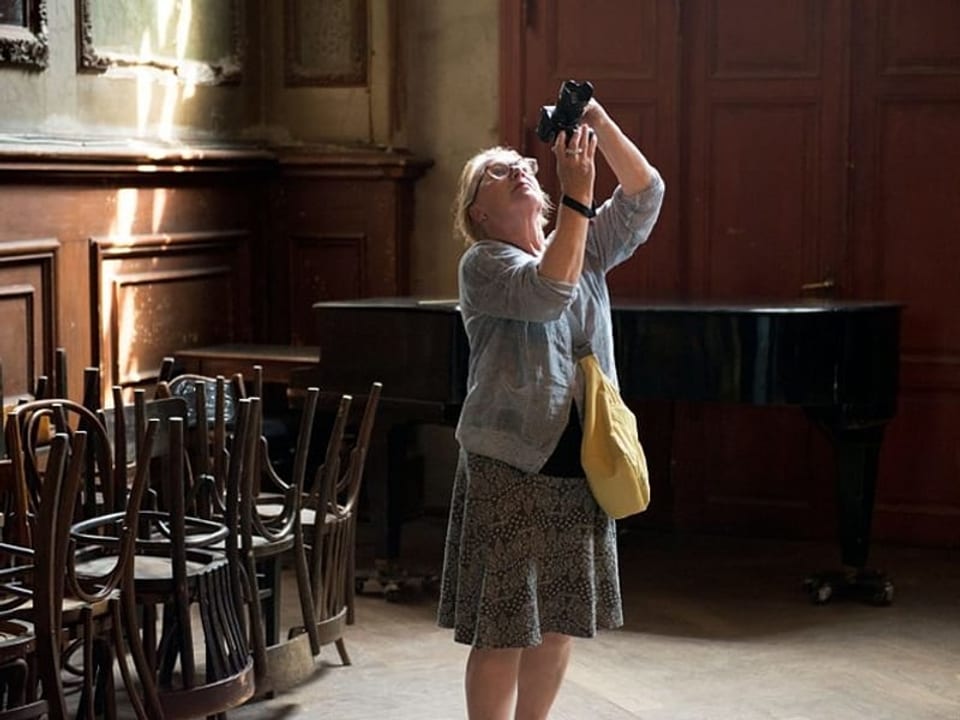 Eine Frau mit einer Kamera in der Hand steht in einem Raum mit Stühlen an den Wänden. 
