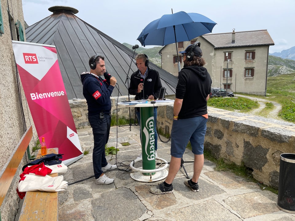 Interviewsituation drei Männer, ein Tisch und im Hintergrund ein Haus. 