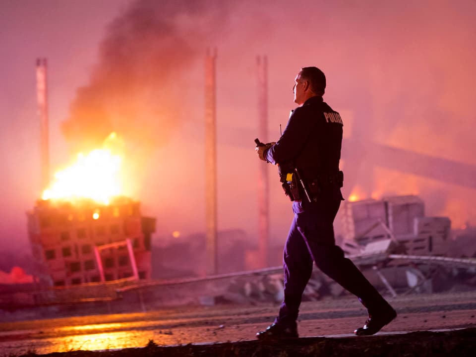 Ein Polizist vor einem brennenden Gebäude, auf einer verwüsteten Strasse. 