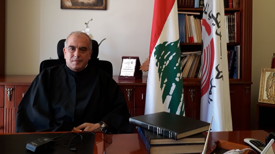 Pater Abdo Abou Kassm, ein älterer Mann, sitz an seinem Schreibtisch, hinter ihm ist die Flagge des Libanons.
