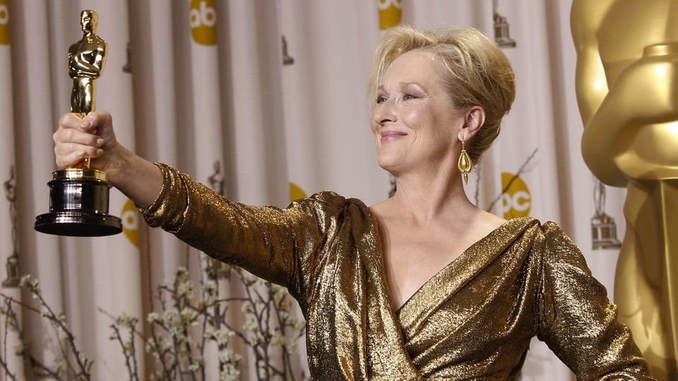 Meryl Streep lächelt und hält mit der rechten Hand den Oscar hoch.