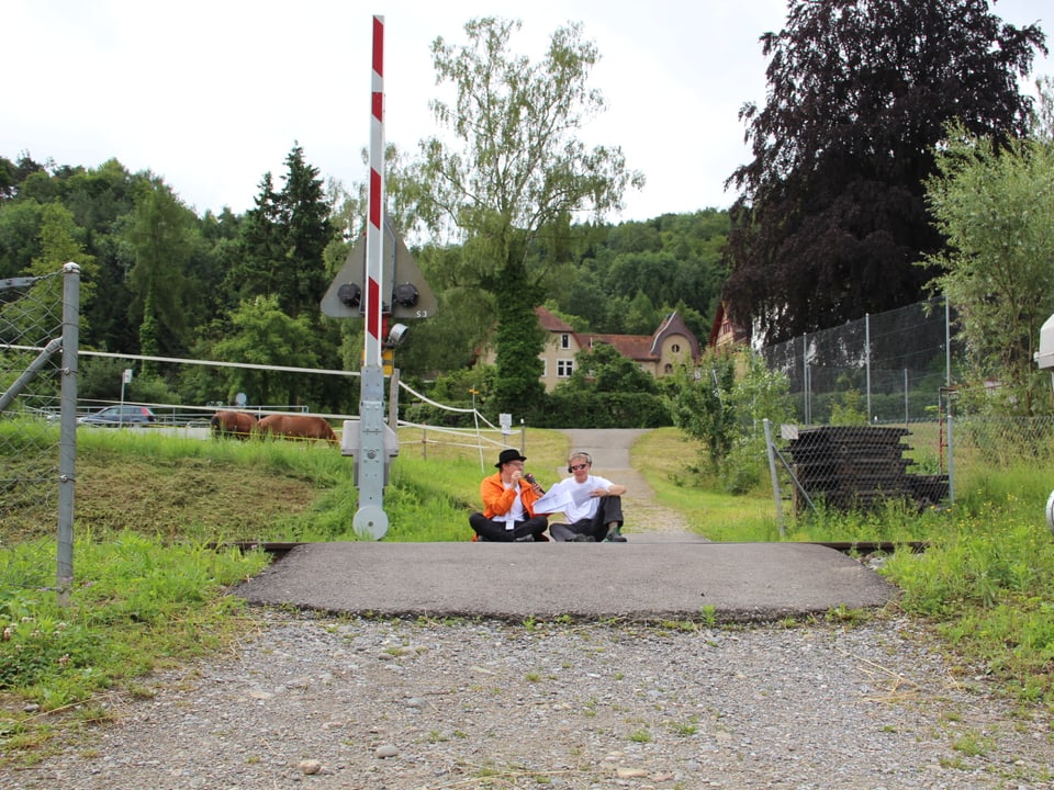 Mundartexperte und Reto Scherrer sitzen am Boden vor einer Bahnbarriere.
