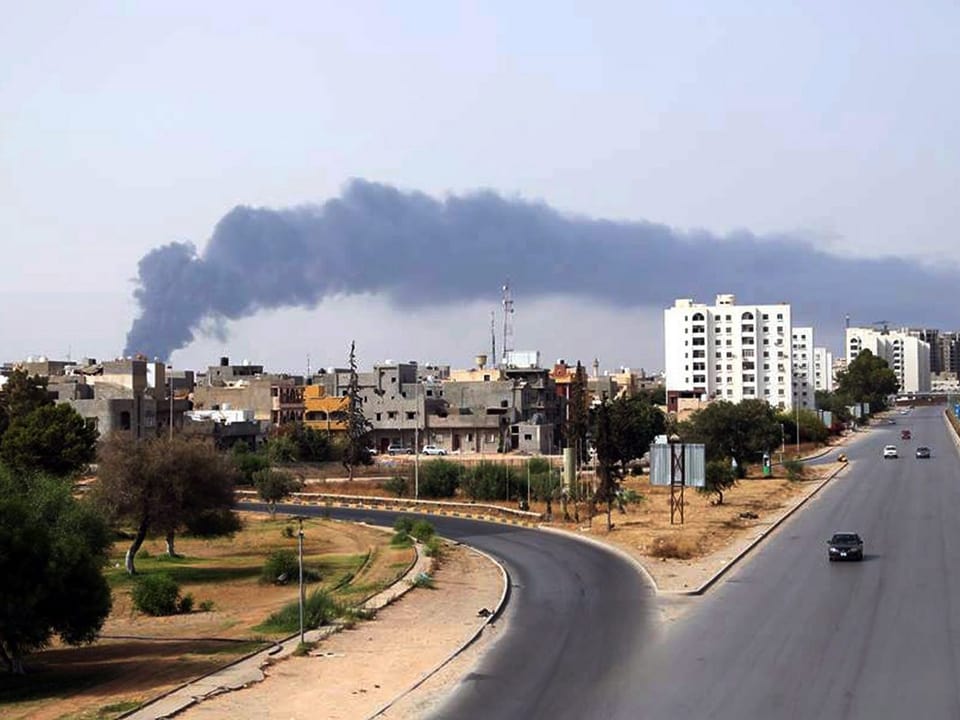 Eine Rauchsäule zieht über die Skyline von Tripolis, im Vorderdund eine fast leere Strasse, die abzweigt.