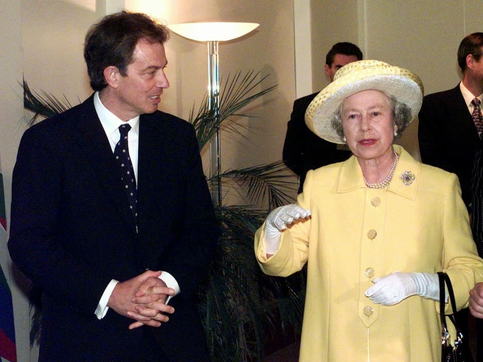 Tony Blair mit Anzug und Krawatte steht neben der Queen, die einen hellgelben Mantel und Hut trägt.