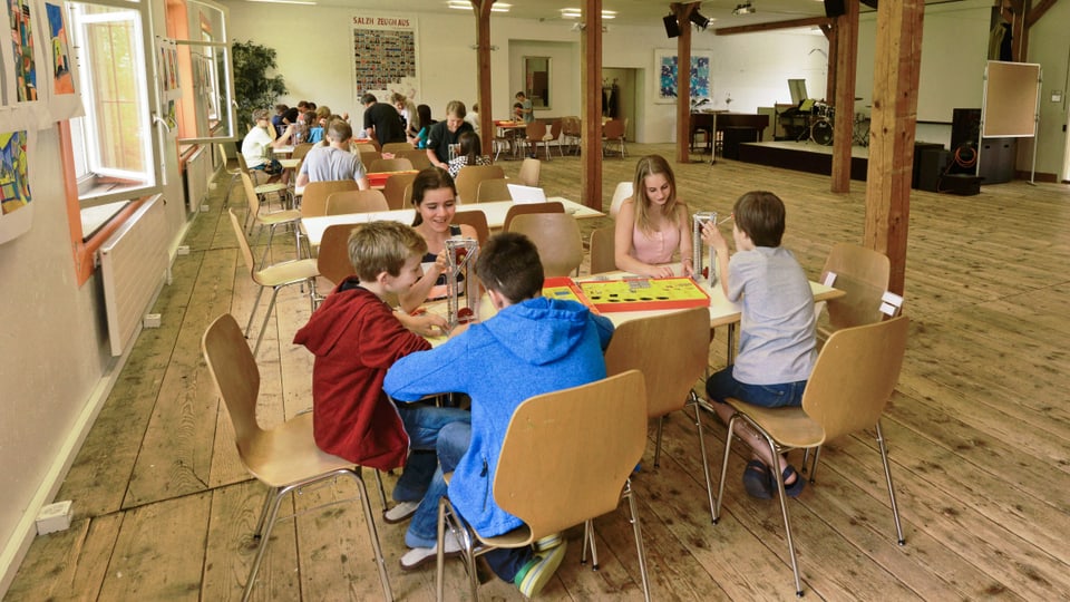 Kinder sitzen in einem grossen Saal an Holztischen