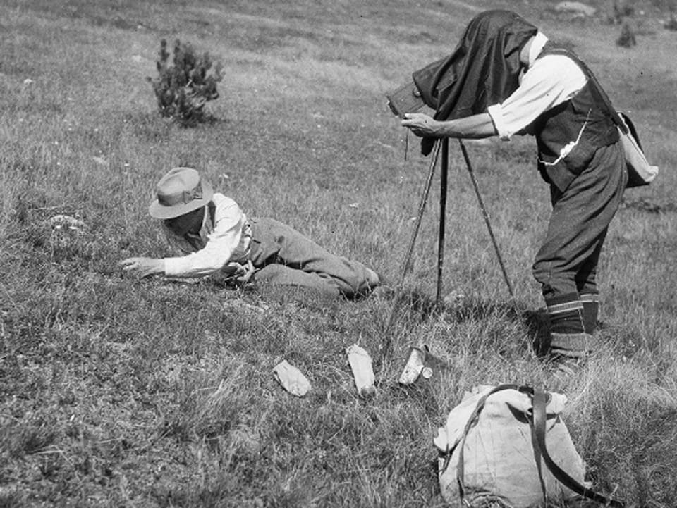 Zwei Männer auf einer Wiese - der eine liegt auf der Wiese und untersucht etwas am Boden, der andere nimmt ein Foto mit einer alten Kamera auf.